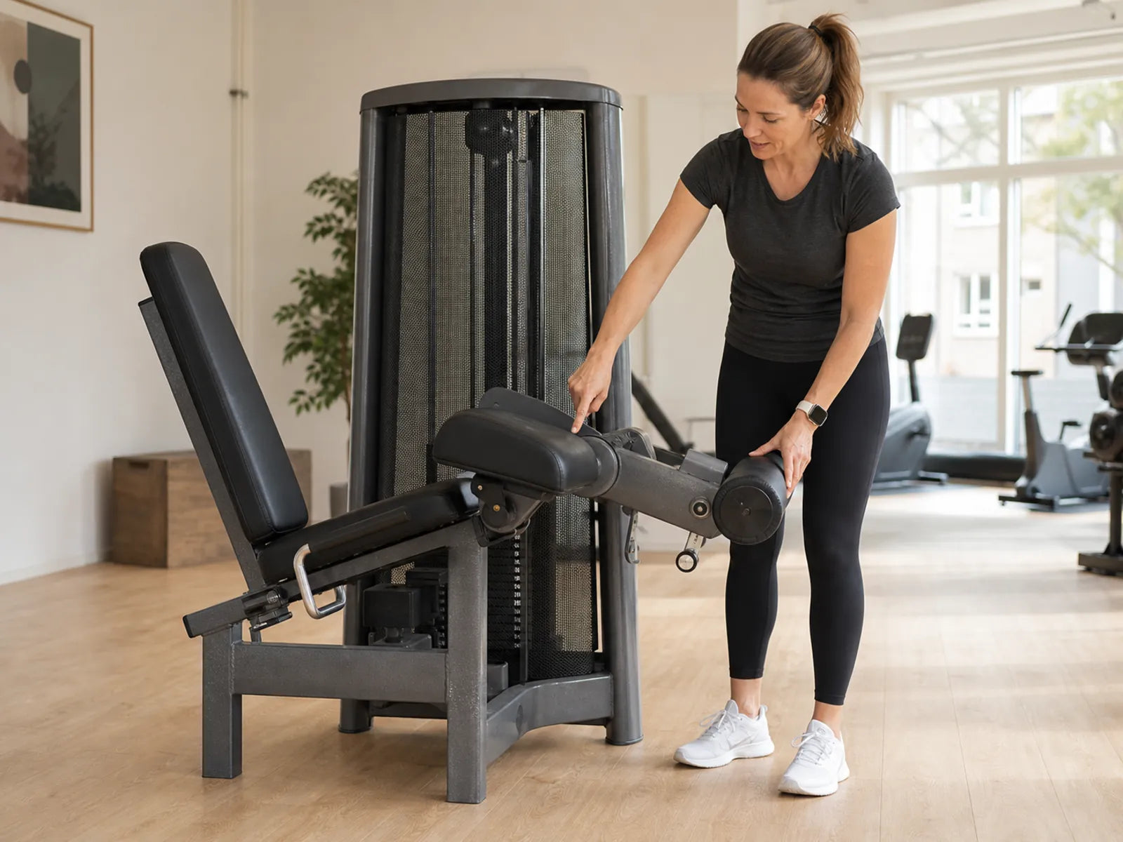Beim Einstellen einer Leg-Curl-Maschine für die Hamstrings kontrolliert eine erwachsene Frau den Drehpunkt und die unbelastete Rolle.